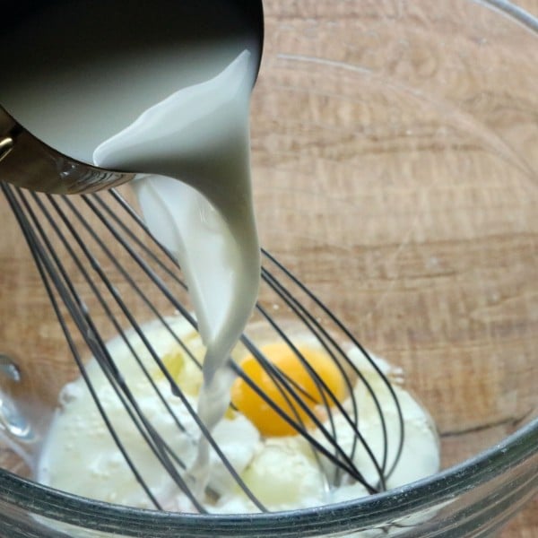 pouring milk over whisks and eggs in a glass mixing bowl on a table