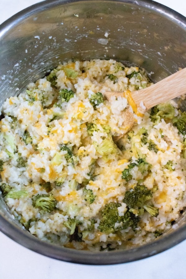 a wooden spoon being used to stir rice, broccoli and cheese in an instant pot on a white counter