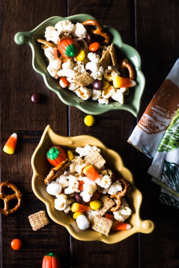 an overhead view of popcorn, mini marshmallows, pretzels, reeses pieces, candy corn and pumpkins, chex cereal in two leaf bowls on a table with more party mix on it 