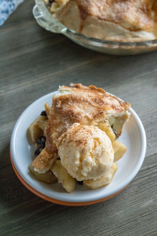 a slice of apple pie topped with a scoop of vanilla ice cream on a white and orange plate on a wood table with the rest of the pie and a linen in the background 