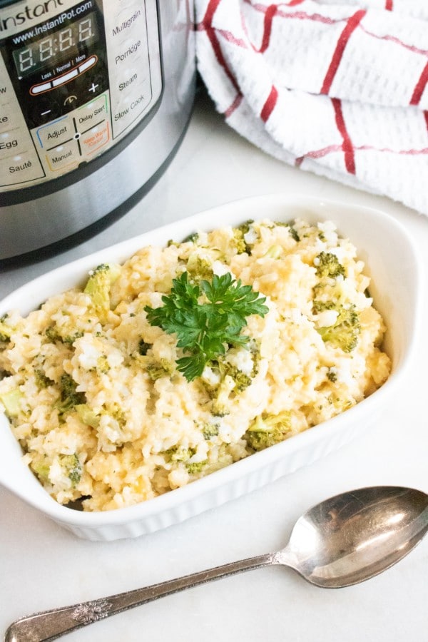 broccoli rice and cheese casserole in a white baking dish on a white counter next to a spoon with an instant pot and red and white cloth in the background with title text reading Instant Pot Broccoli Rice and Cheese Casserole