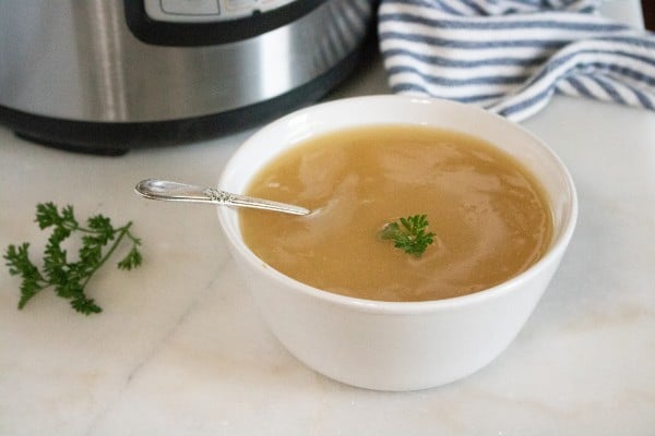 gravy in a white bowl with a spoon in it next to a parsley sprig on the white counter with an instant pot and blue and white linen in the background