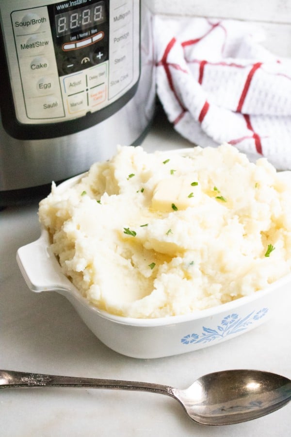 mashed potatoes in a white casserole dish next to a spoon with an instant pot and red and white cloth in the background on a white counter 