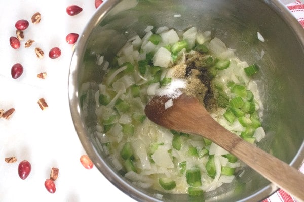 butter, chopped celery and onions, and seasonings being cooked in an instant pot with a wooden spoon with more cranberries and nuts next to it on the white counter and also a red and white linen