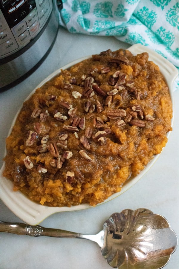 an overhead view of sweet potato casserole tipped with pecans in a baking dish on a white counter next to a serving spoon with an instant pot and green and white cloth in the background 