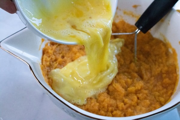 eggs being poured from a white bowl into a white bowl of mashed sweet potatoes