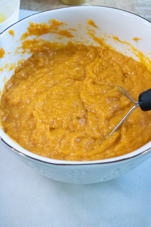mashed sweet potato in a white bowl with a masher in it on a white counter