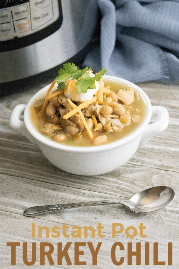 turkey chili topped with sour cream, shredded cheese and parsley, in a white bowl next to a spoon, instant pot and blue cloth on a wood table with title text reading Instant Pot Turkey Chili