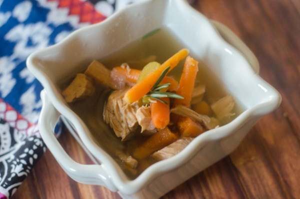 turkey soup topped with sliced carrots in a white bowl on a wood table with a multi-colored cloth in the background