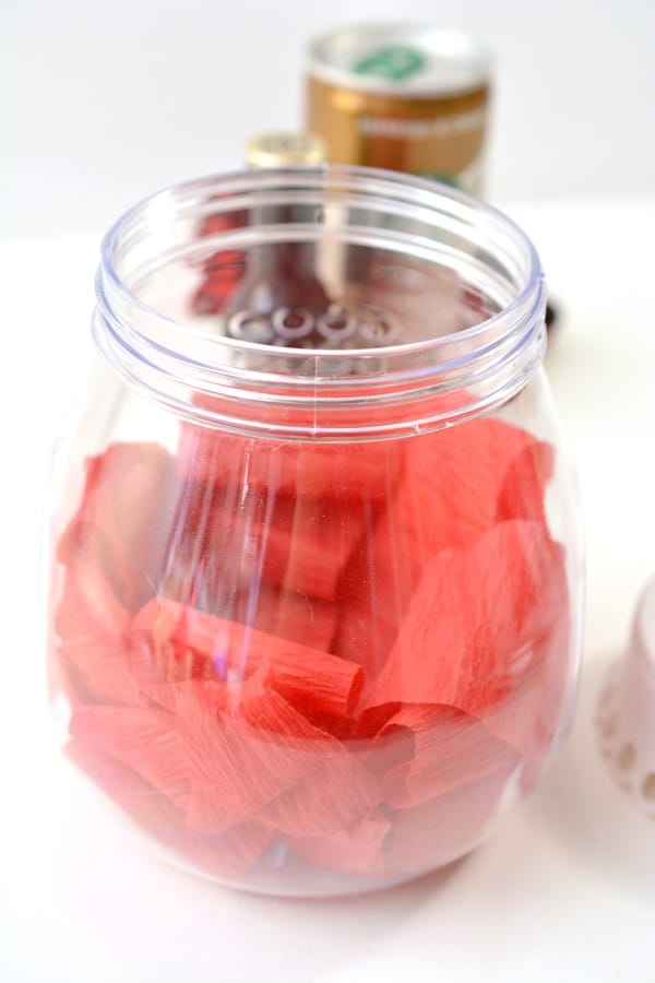 a glass jar with red crepe paper in it with a can of coffee and a bottle of liquer in the background