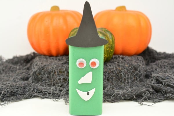 a juice box decorated with green, white and black paper to look like a witch on a white table with black netting and three pumpkins in the background