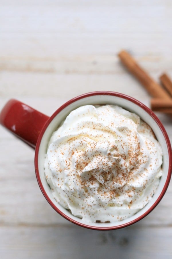 an overhead view of hot chocolate in a red mug topped with whipped cream with cinnamon sticks next to it on a table