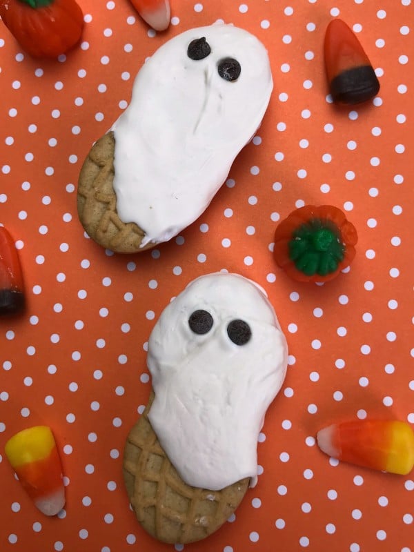 nutter butters decorated with white frosting and mini chocolate chips to look like ghosts on an orange and white polka dot background next to candy pumpkin and candy corn 