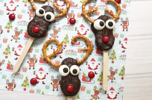 nutter butter cookies covered in chocolate with candy eyes, a red M&M nose, pretzel antlers on a popsicle stick on a Christmas paper background