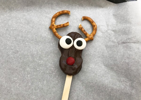 a nutter butter cookie covered in chocolate with candy eyes, a red M&M nose, pretzel antlers on a popsicle stick on a parchment paper