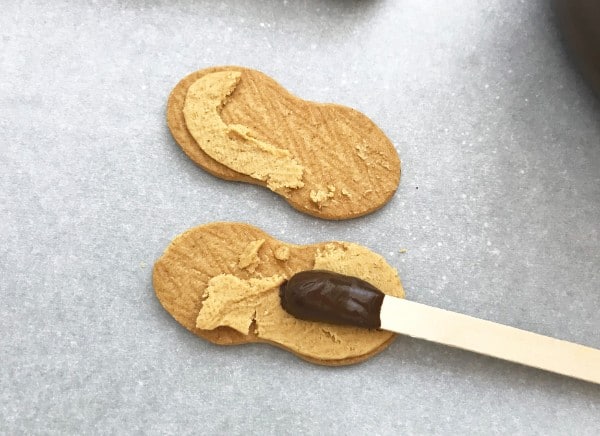 a nutter butter cookie broken in half with a popsicle stick with melted chocolate on one end on one half of the cookie on a gray background