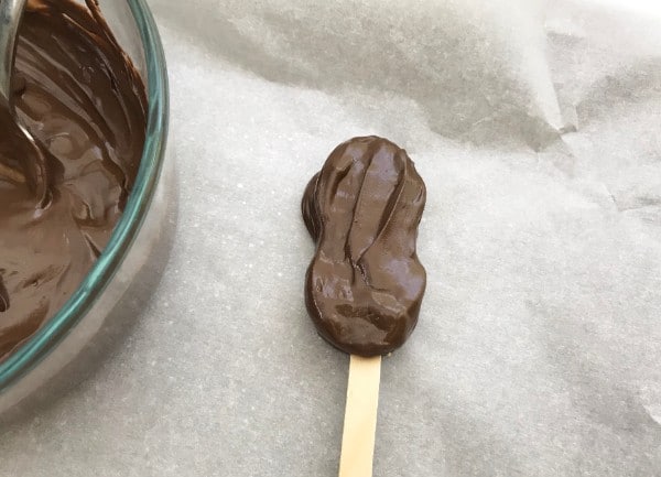 nutter butter cookie covered in melted chocolate with a popsicle stick stuck in it next to a glass bowl of melted chocolate on a gray background