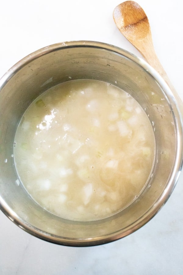 overhead view of rice, chicken broth, salt, garlic and onions in an instant pot next to a wooden spoon on a white counter