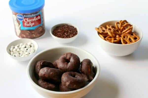 a can of chocolate frosting, white bowls of candy eyes, chocolate sprinkles, pretzels and mini chocolate donuts on a white table