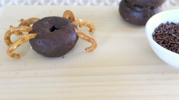 a mini chocolate donut with broken pretzels stuck in the sides to look like spider legs on a brown cutting board with another donut and a white bowl of chocolate sprinkles int the background