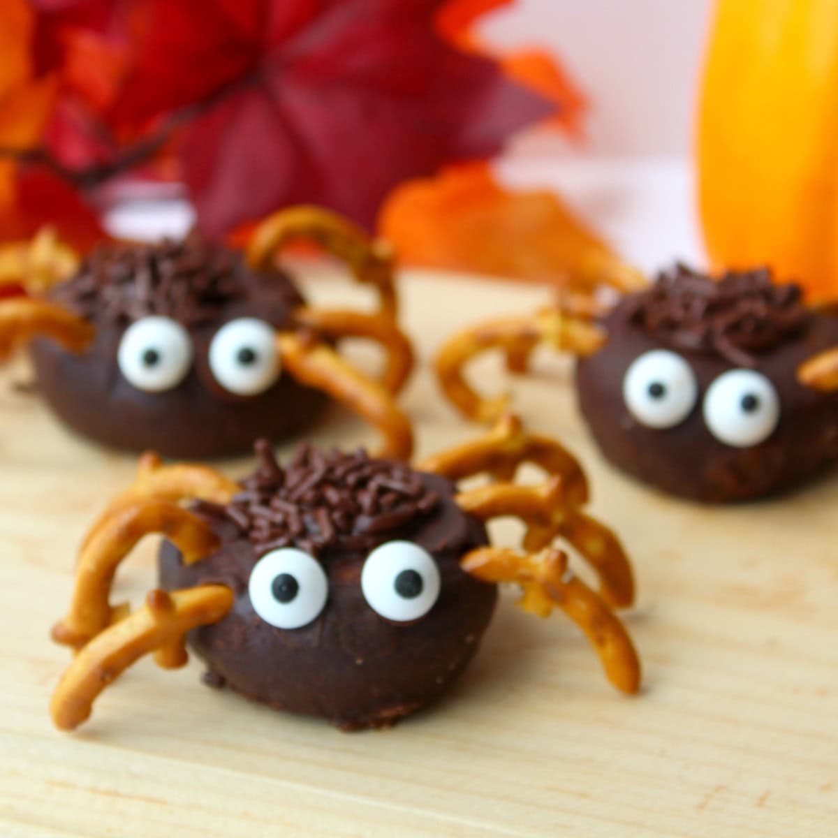 Three mini donut spider treats made of chocolate, candy eyes, and pretzel legs on a wooden surface.