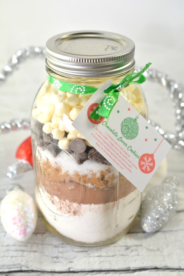 layered cookie mix in a glass jar with a card tied on it with a green ribbon with text reading chocolate lovers cookies on it on a wood table with silver decorations around it