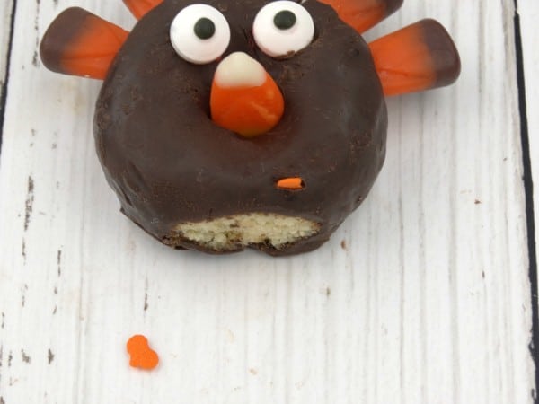 a mini chocolate donut with candy corn stick in the top of it, with candy eyes and a candy corn nose, and leaf sprinkles as feet on a table