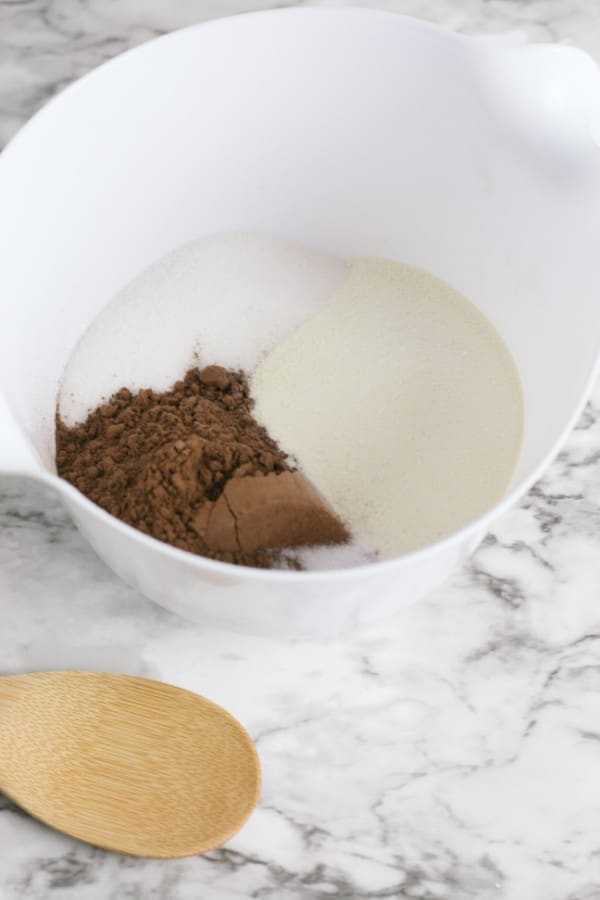 sugar, baking cocoa, powdered milk, and a pinch of salt in a white mixing bowl on a counter next to a wooden spoon
