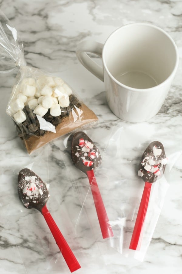 a bag of hot cocoa mix, marshmallows, and chocolate chips next to a mug of hot chocolate and red plastic spoons coated with melted chocolate and crushed peppermints all on a counter