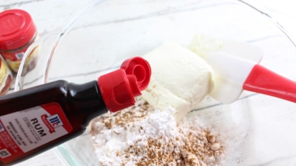 rum extract being poured into cream cheese, powdered sugar, cinnamon, nutmeg in a glass mixing bow
