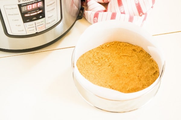 graham cracker crust in a paper lined springform pan with an instant pot in the background, all on a white table