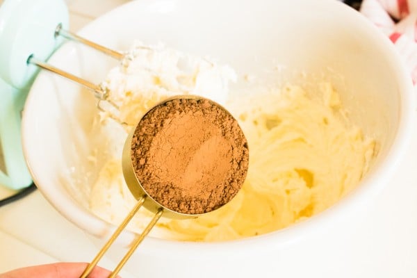 a measuring cup of cocoa powder above the mixing bowl of cream cheese and sugar