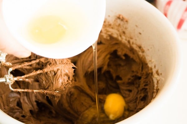 eggs being poured from a bowl into a mixing bowl of chocolate, cream cheese and sugar
