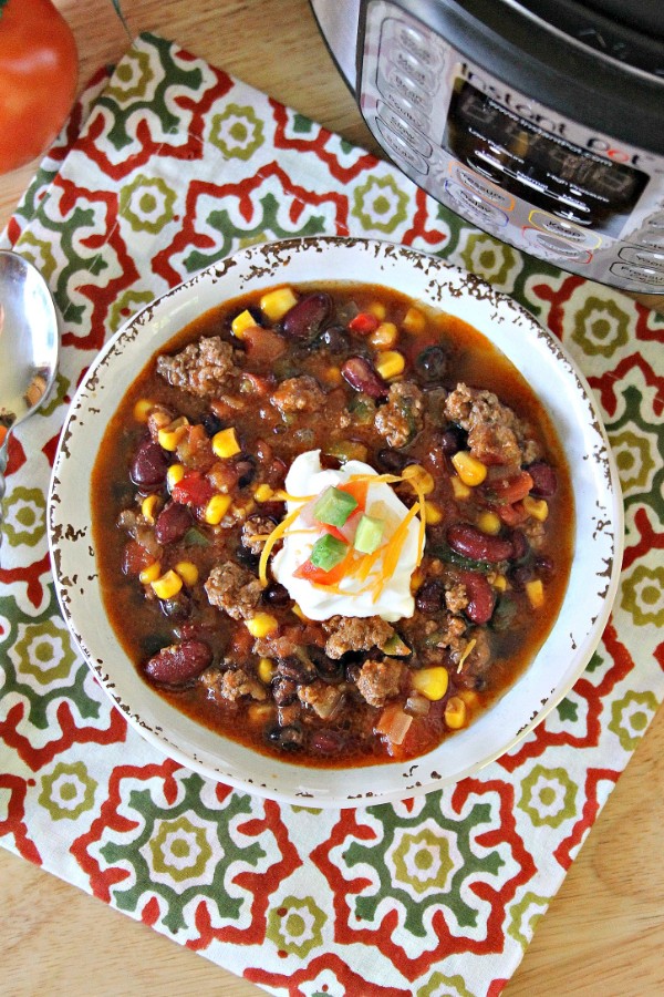 an overhead view of taco soup in a white bowl topped with sour cream on a flower linen on a brown table with a spoon, tomato and instant pot in the background