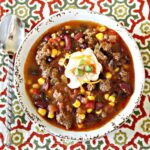 A bowl of savory chili, reminiscent of a hearty Instant Pot taco soup, with ground meat, corn, and kidney beans. Topped with sour cream and herbs on a patterned tablecloth, a spoon rests beside this delightful dish.