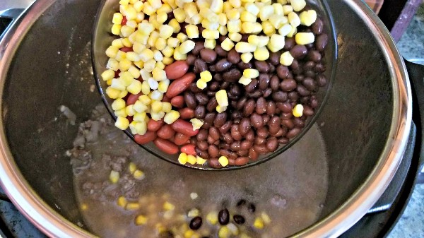black beans, kidney beans, and corn being added from a glass bowl into an instant pot with ground beef and broth