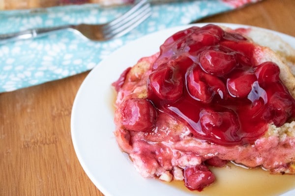 french toast topped with cherries on a white plate with a fork on a blue cloth in the background 