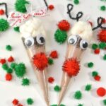 Two reindeer-themed hot chocolate gift bags feature googly eyes, red pom-pom noses, and antlers, all surrounded by festive red and green pom-poms on a pristine white surface.