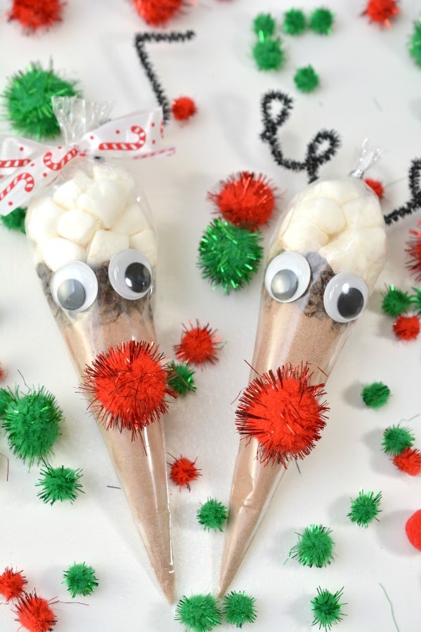 hot cocoa in a plastic bag shaped like a cone with a red pompom and googly eyes glued on it with more red and green pompoms on the table around them