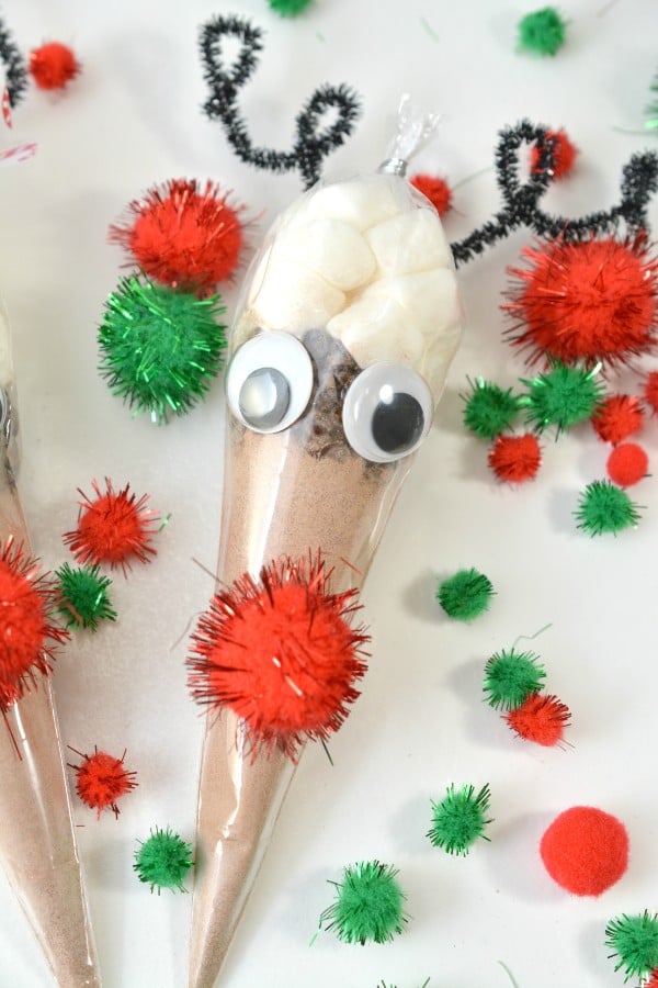 hot cocoa in a plastic bag shaped like a cone with a red pompom and googly eyes glued on it with more red and green pompoms on the table around it