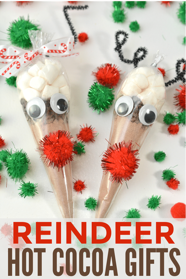 hot cocoa in a plastic bag shaped like a cone with a red pompom and googly eyes glued on it with more red and green pompoms on the table around them with title text reading Reindeer Hot Chocolate Gifts