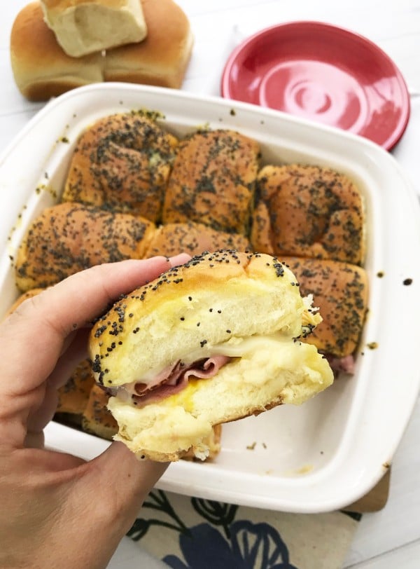 a hand holding a ham and cheese hawaiian roll sliders topped with sauce and poppy seeds  above  a white dish filled with more sliders with more rolls and a saucer in the background
