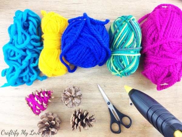 five colors of yarn, pine cones, scissors, and a glue stick on a table