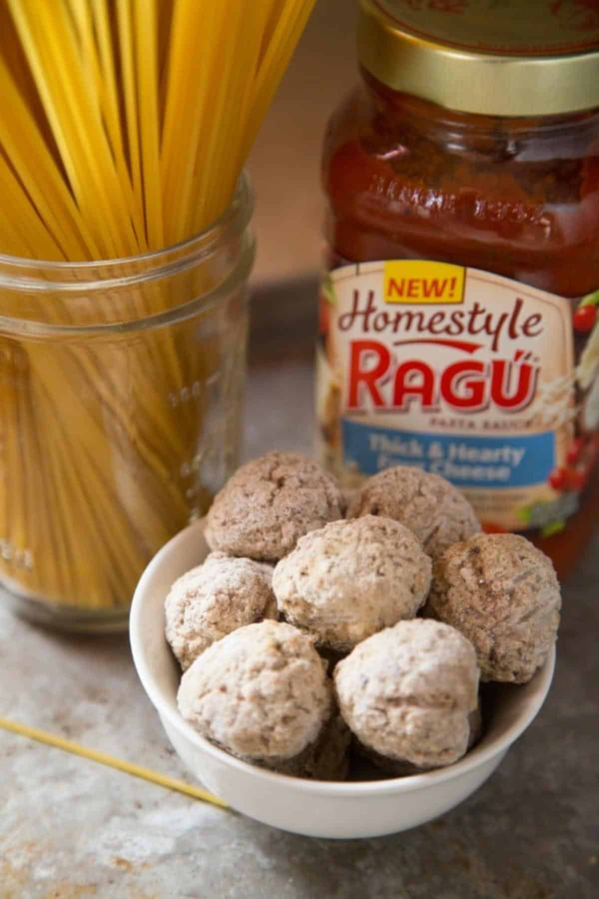 A jar full of spaghetti noodles, a white bowl of frozen meatballs, a jar of ragu pasta sauce.