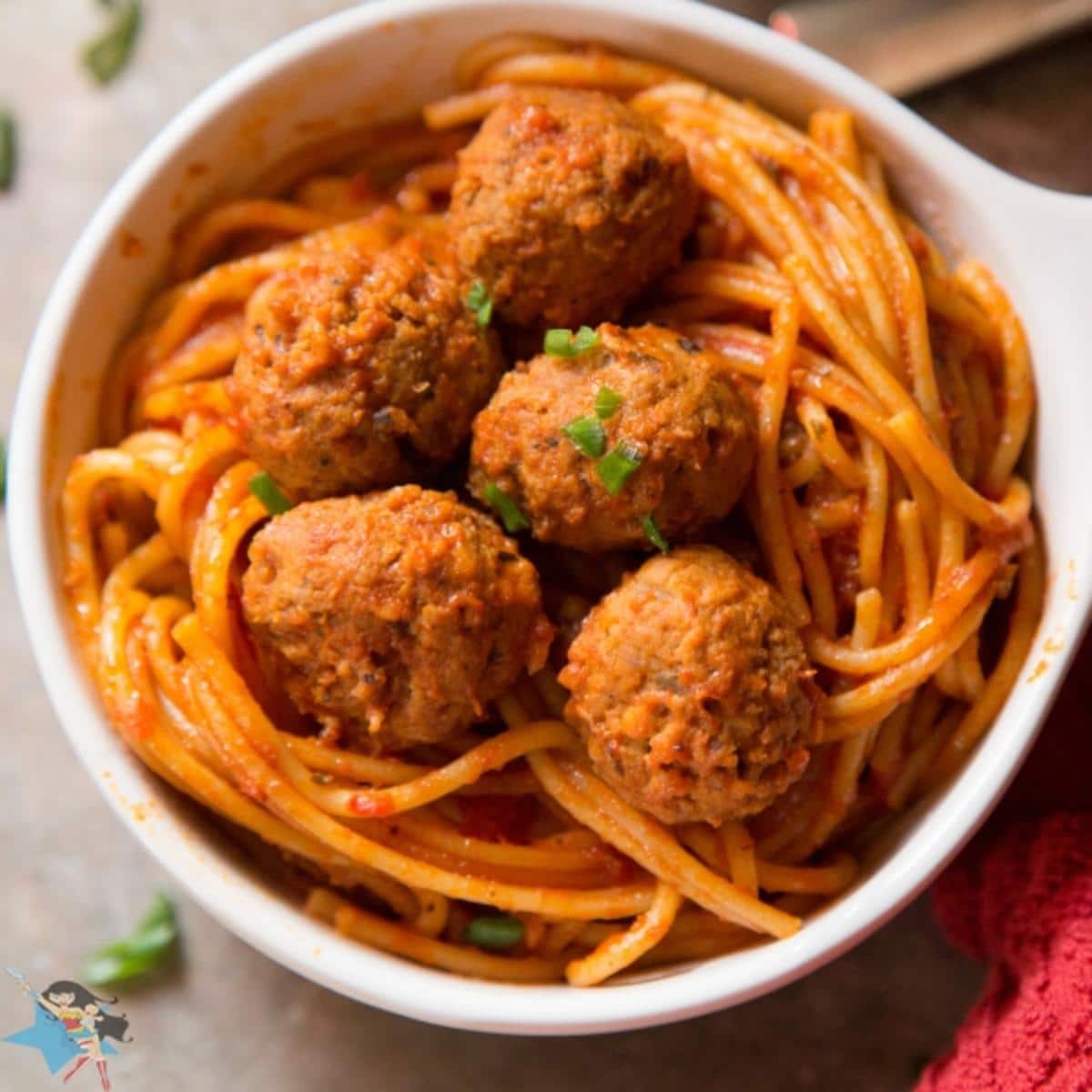 A white bowl of Spaghetti and Meatballs on a table with a red cloth next to it.