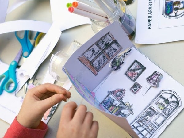  a child putting together a printable doll house with scissors and a glass jar of markers on the table 
