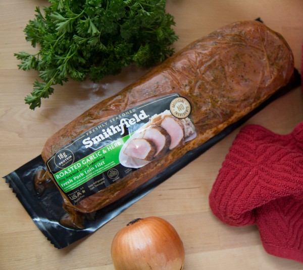 roasted garlic & herb pork loin filet in a package on a table next to an onion, red cloth and bunch of parsley