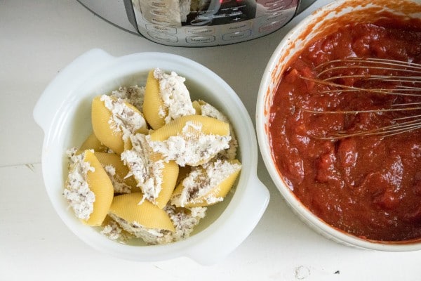 pasta shells stuffed with sausage and cheese in a white bowl next to a white bowl of pasta sauce with a whisk in it with an instant pot in the background