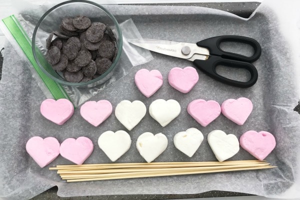 heart shaped marshmallows, chocolate candy melts in a glass bowl, scissors, wooden skewers, plastic bag all on wax paper on a baking sheet