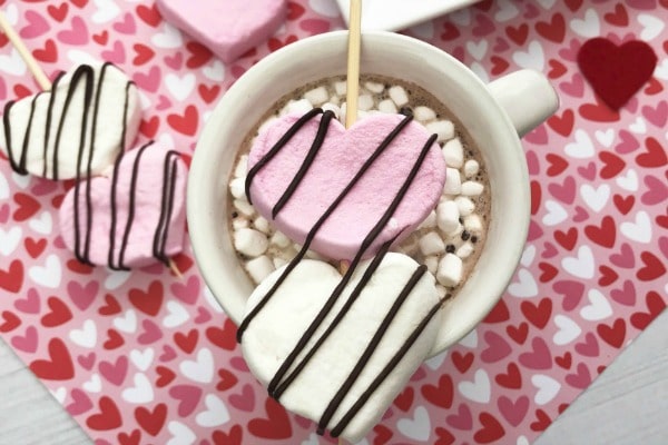 heart shaped marshmallows on wooden skewers on the top of a mug of hot cocoa topped with mini marshmallows all on a heart linen with more marshmallows next to it